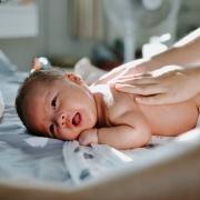 white newborn baby lying belly down with two hands (belonging a person not in the frame) on the baby's back