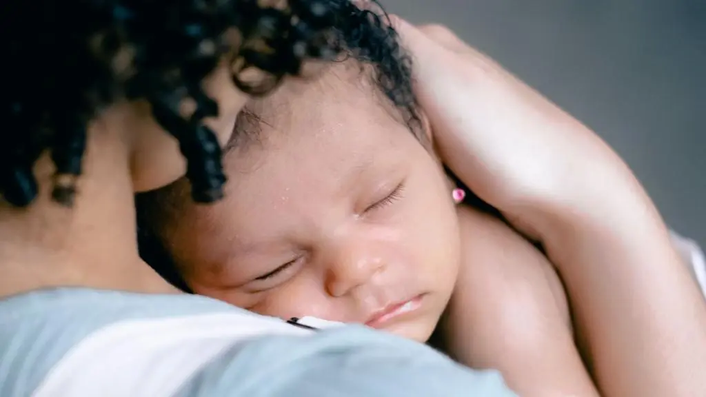 A baby sleeps peacefully on their curly-haired caregiver's shoulder.