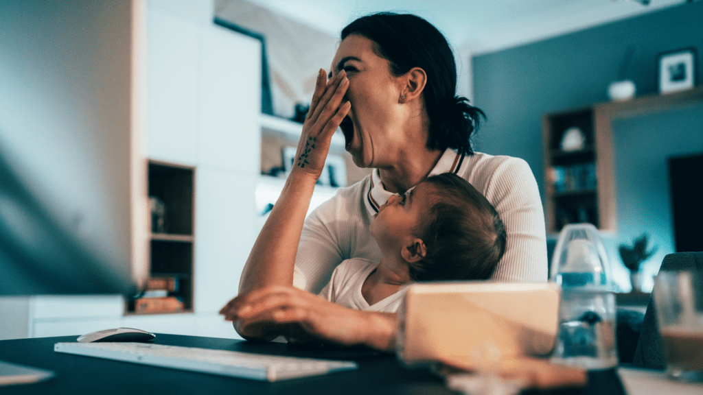 A tired parent yawns as their baby watches while they work from home.