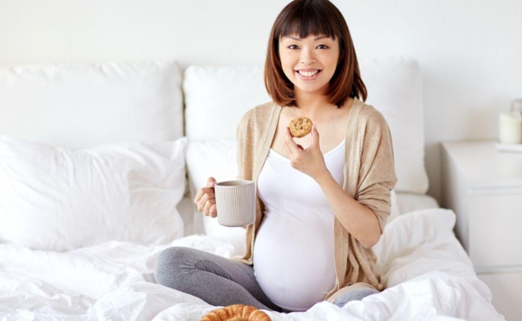Pregnant person sitting on bed, smiling, enjoying cookies. Feeling prepared by childbirth education and birth doula support in Baltimore