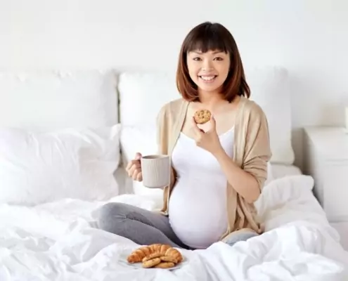 Pregnant person sitting on bed, smiling, enjoying cookies. Feeling prepared by childbirth education and birth doula support in Baltimore
