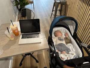 A baby in a Mockingbird Stroller with Bassinet sleeps next to a table holding two drinks and a laptop. Mother's Day Gift Guide